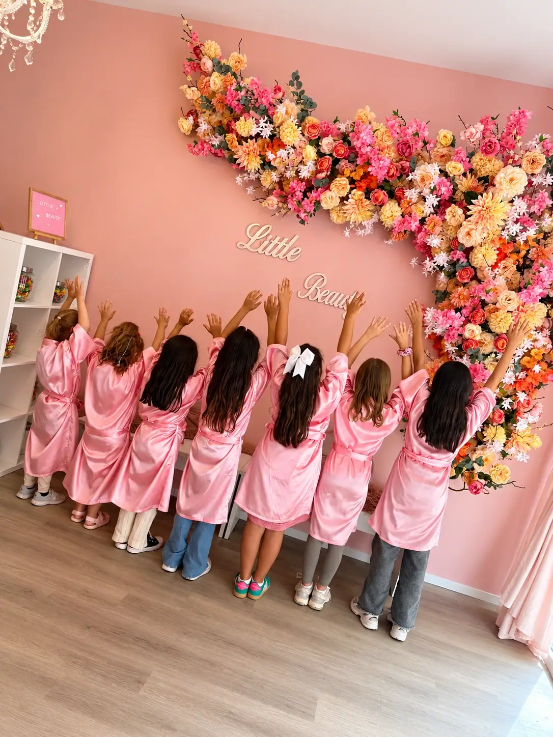 Sieben Mädchen in rosa Spa-Roben vor der Blumenwand neben dem Candy-Regal bei Little Beauty
