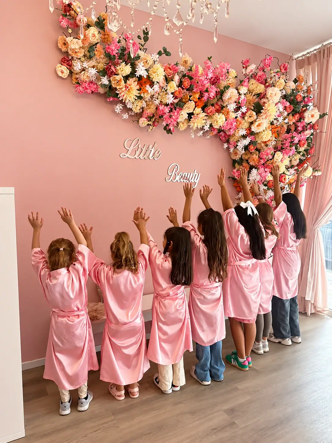 Mädchen in rosa Spa-Roben strecken die Arme hoch vor der Blumenwand im Little Beauty Salon in Düsseldorf