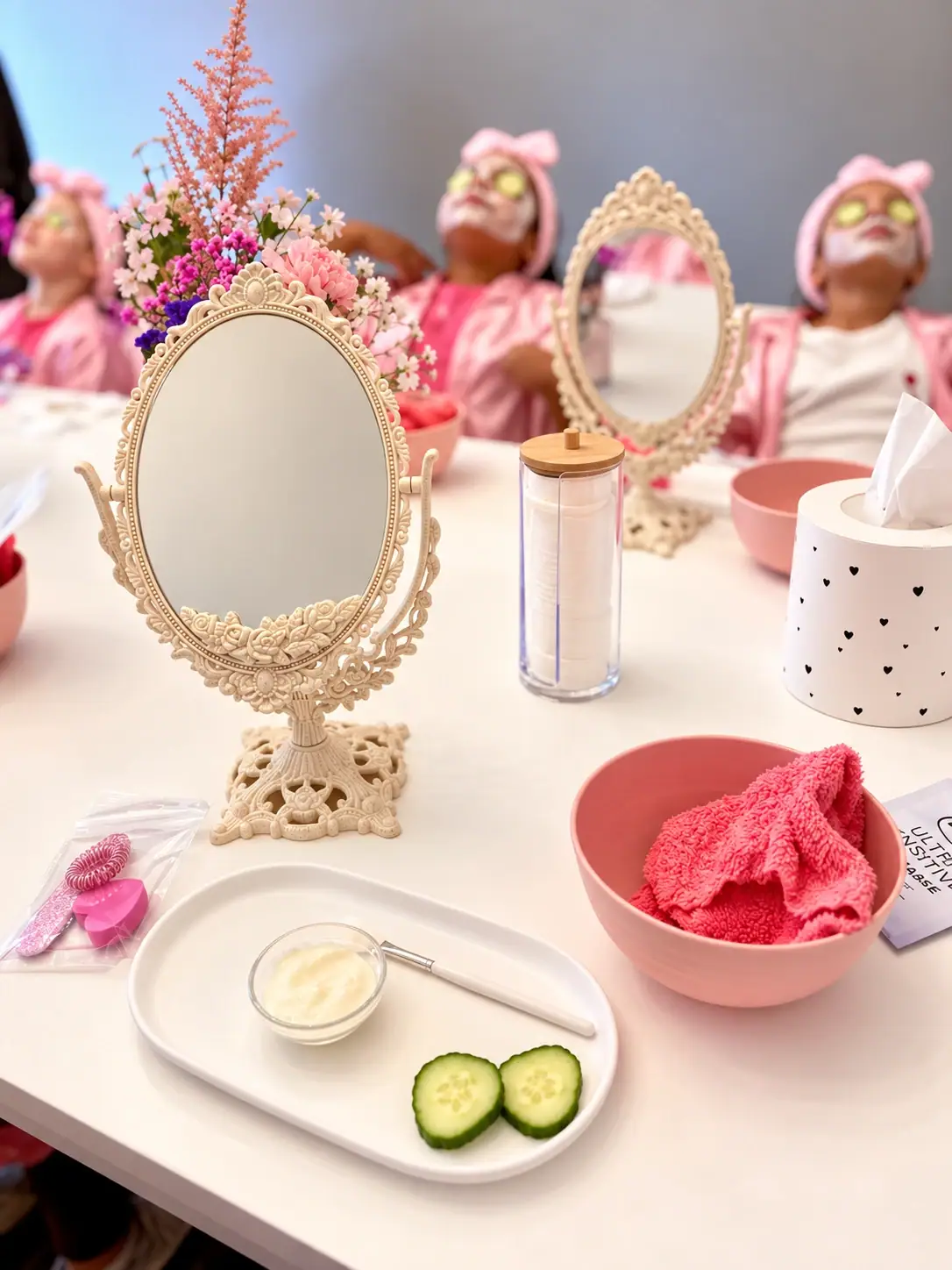 Gedeckter Spa-Tisch mit Vintage-Spiegeln, Gurkenscheiben und Gesichtscreme, dahinter Kinder mit Gesichtsmaske