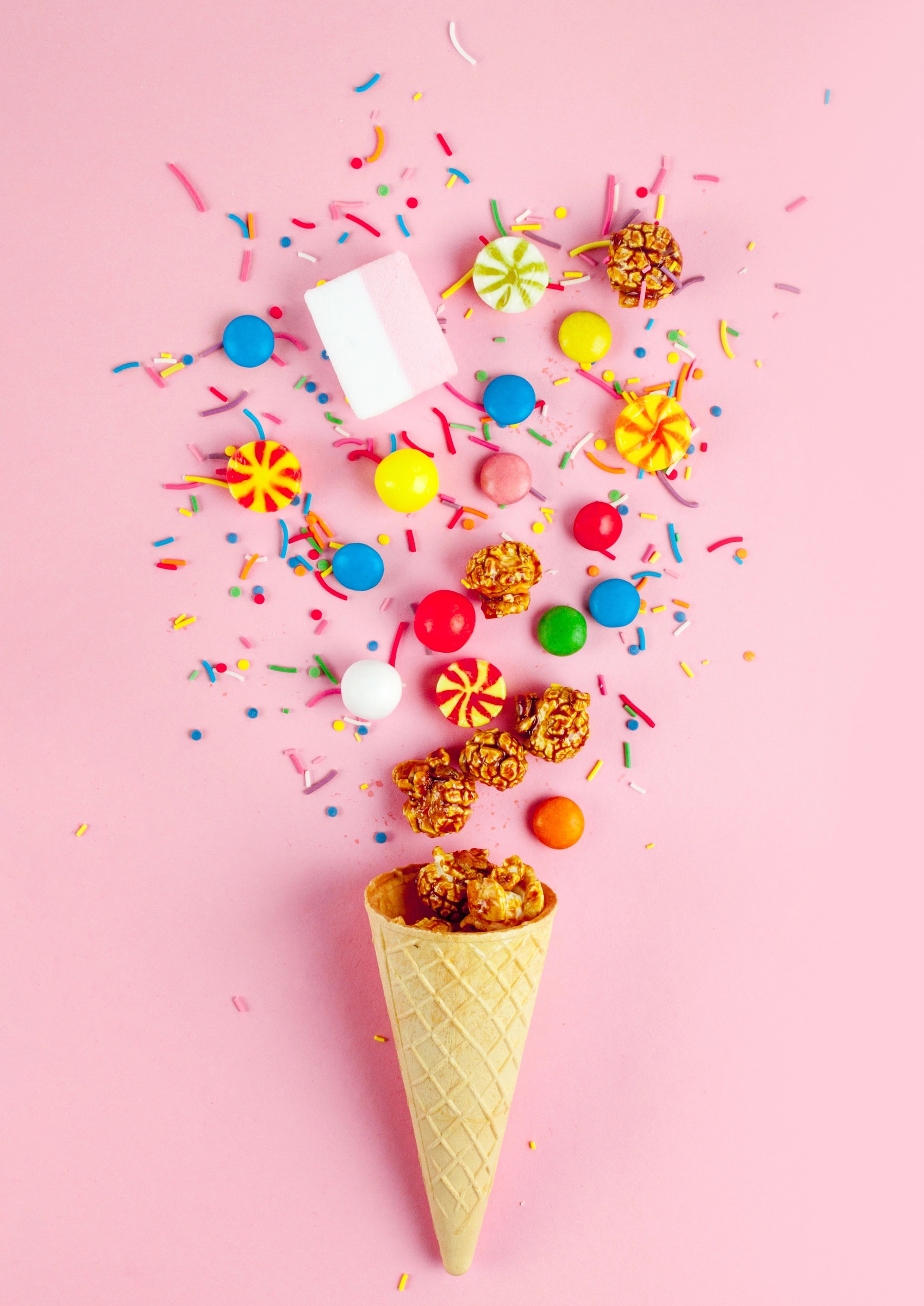 Bunte Süßigkeiten, Popcorn und Zuckerstreusel in einer Waffeltüte auf rosa Hintergrund – kreative Candy-Bar-Idee für Kindergeburtstag und Spa-Party für Mädchen.