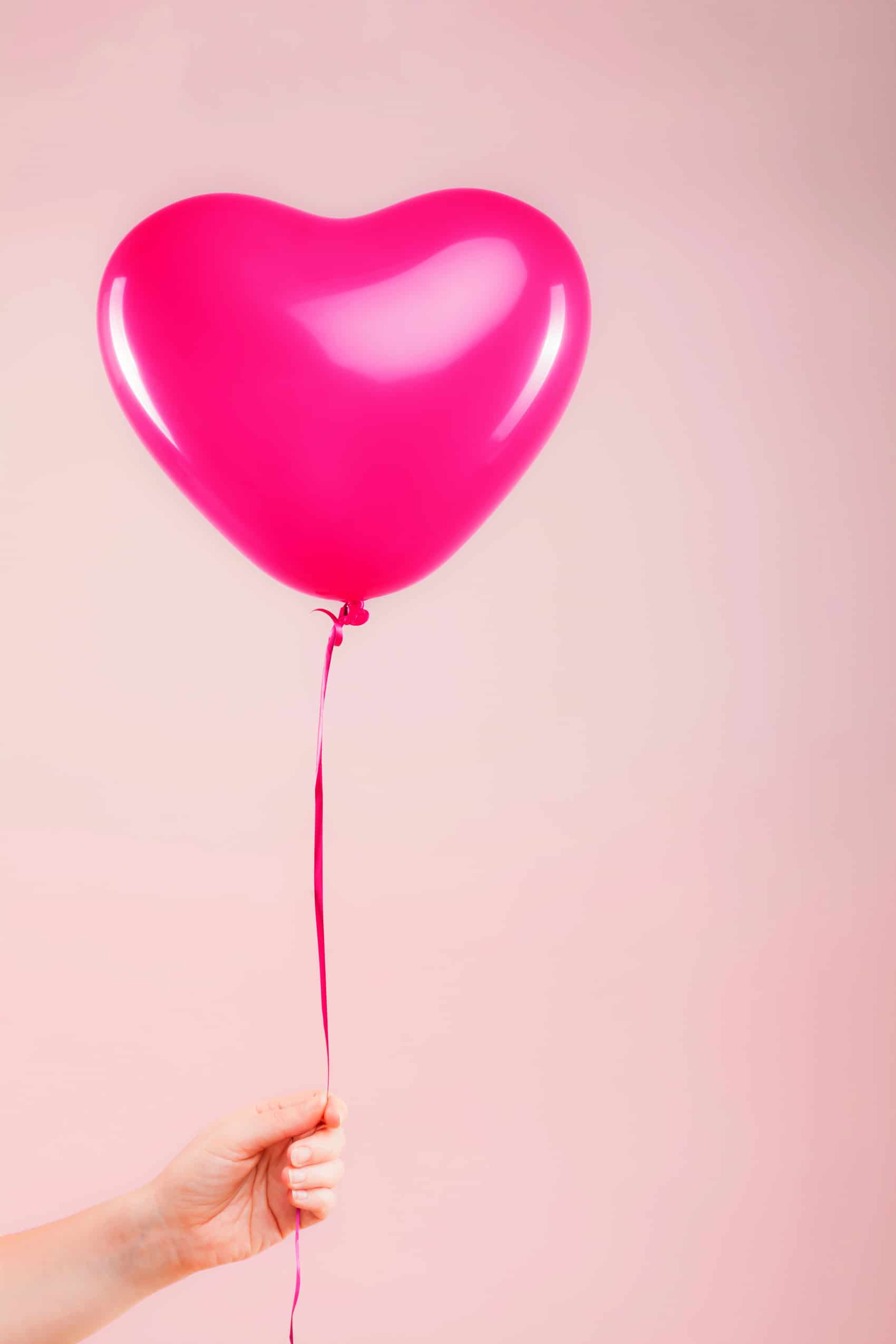 Hand hält rosa Herzluftballon vor rosa Hintergrund
